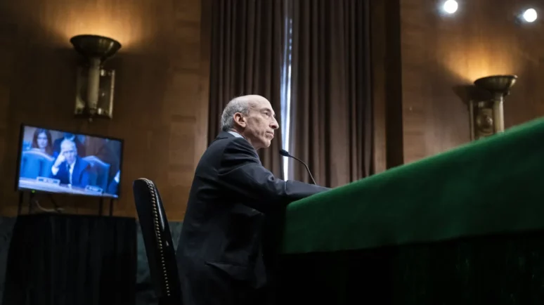 SEC Chairman Gary Gensler sitting at a hearing, TV screen behind him.